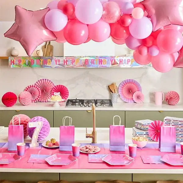 Pink birthday party table with a balloon arch, star balloons, a lit number 7 decoration, gift bags, party tableware, and birthday decorations.